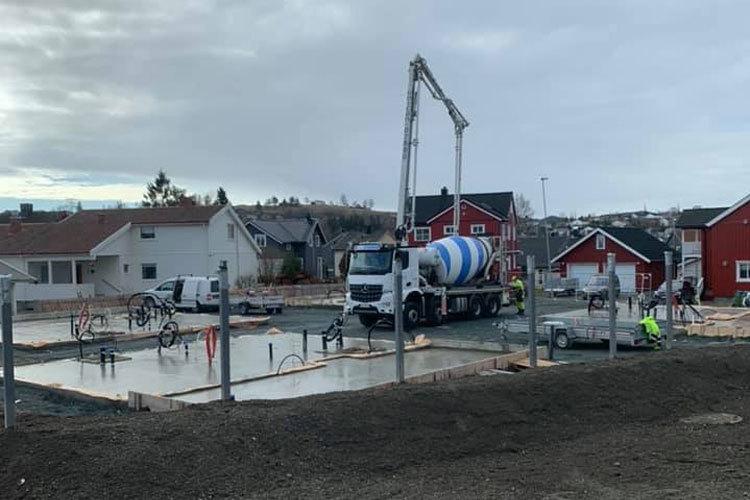 Betongbil leverer betong til byggeplass med hus i bakgrunnen, arbeidere jobber med fundamentet.