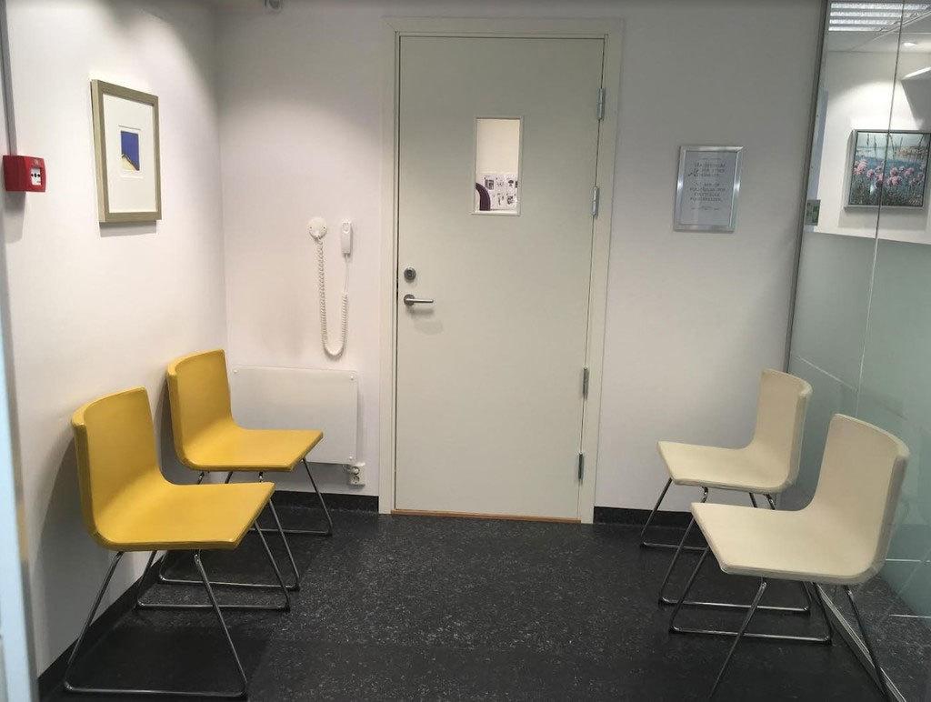Venterom med fire gule stoler, en dør og en telefon på veggen.