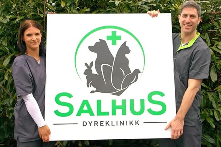To personer holder opp et skilt med logoen til Salhus Dyreklinikk, omgitt av grønne planter.