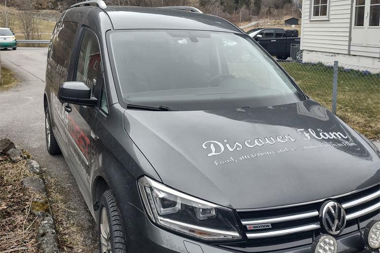 En grå Volkswagen varebil med tekst på siden, parkert ved en husvegg i et boligområde.