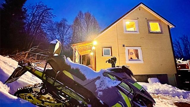 En snøscooter står i snøen foran et gult hus med opplyste vinduer om kvelden.