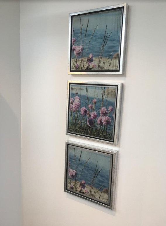Tre innrammede malerier av blomster og hav, hengt på en hvit vegg.