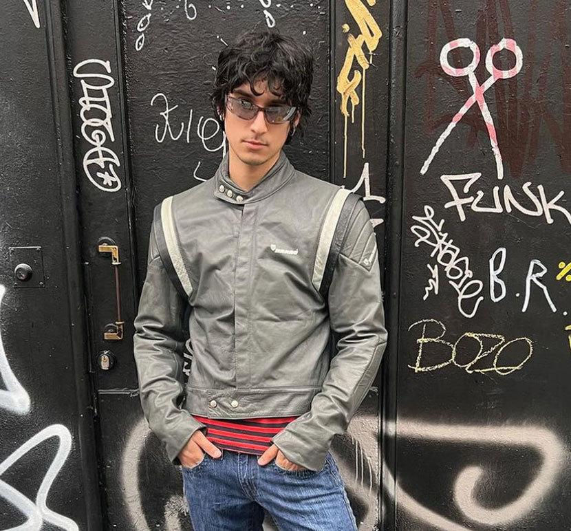 Ung mann med briller iført en grå skinnjakke, står foran en vegg dekket av graffiti.