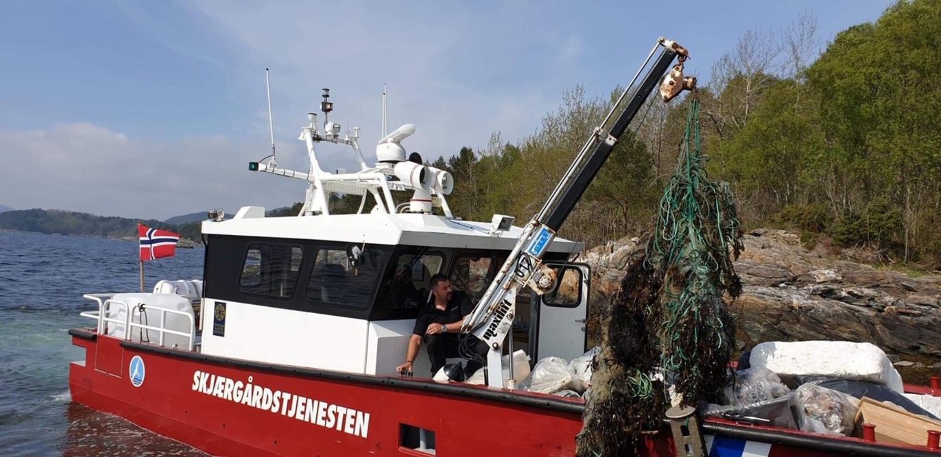 Fiskebåt med norsk flagg som henter opp garn fra sjøen, omringet av grønn natur og klart vann.