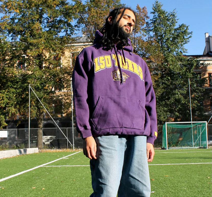 En mann med dreadlocks står på en fotballbane iført en lilla hettegenser og jeans.