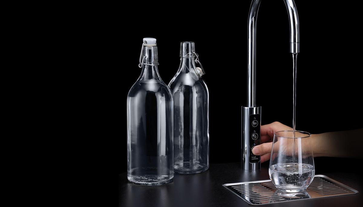 En hånd fyller et glass med vann fra en moderne kran, med to glassflasker i bakgrunnen.