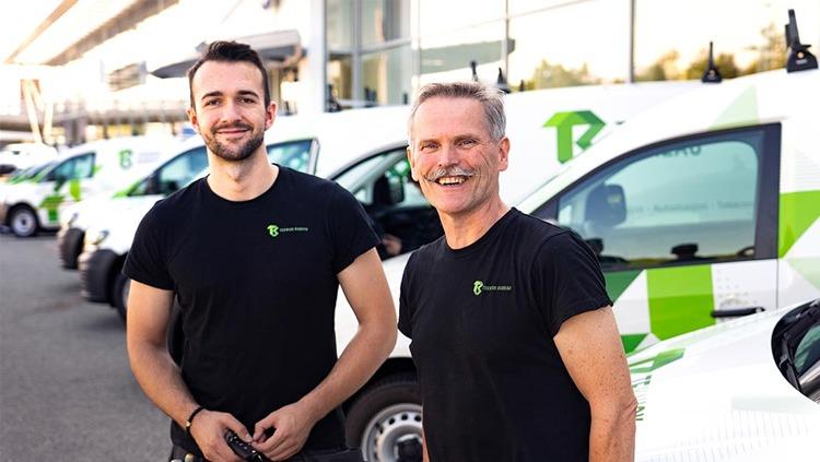 To personer i svarte t-skjorter står foran flere hvite varebiler med grønt logo.
