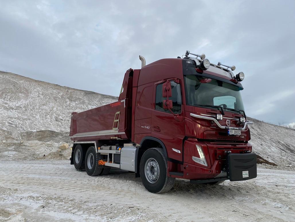 En rød lastebil med tippvogn står på snødekt underlag, omgitt av en skråning.