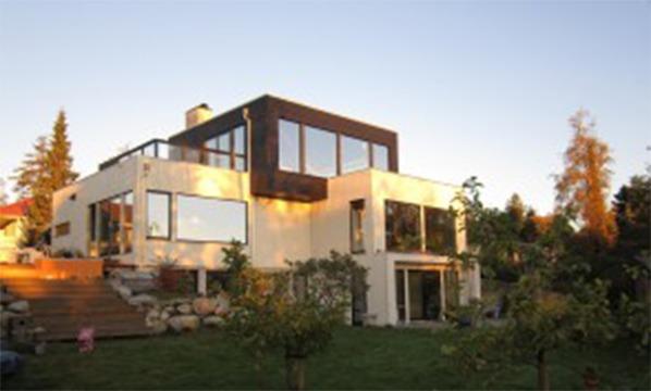 Moderne hus med store vinduer og terrasse, omgitt av trær og hage i solnedgang.