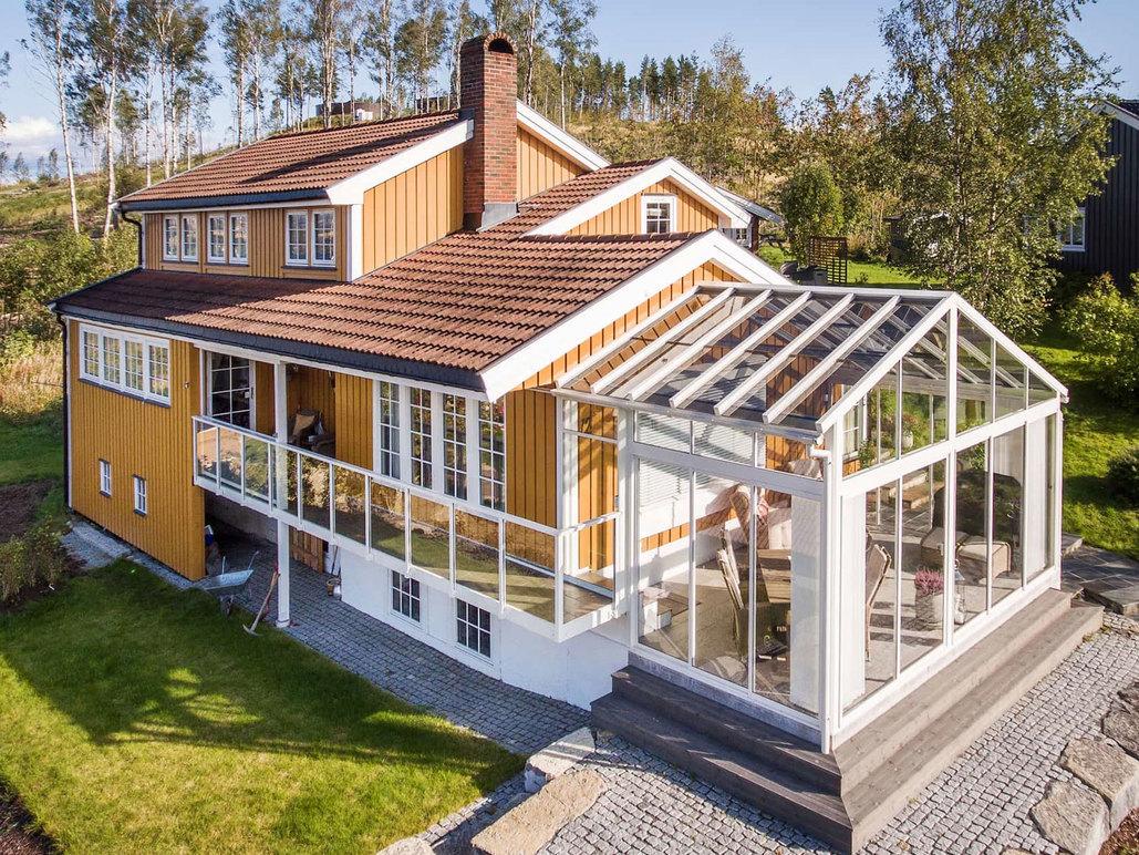 En vakker, moderne hytte med trepanel og glassveranda, omringet av grønt landskap og trær.