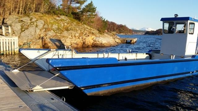 Båt med blått skrog ligger ved kaien, med fjell og trær i bakgrunnen under klar himmel.