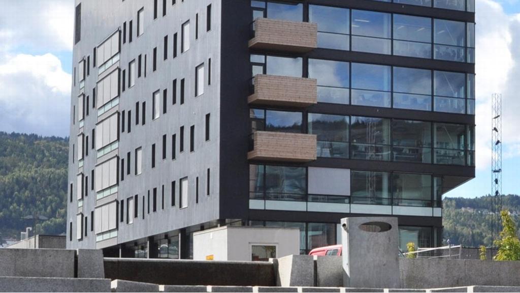Moderne kontorbygg med store vinduer og trepanel, omgitt av grønt landskap og blå himmel.