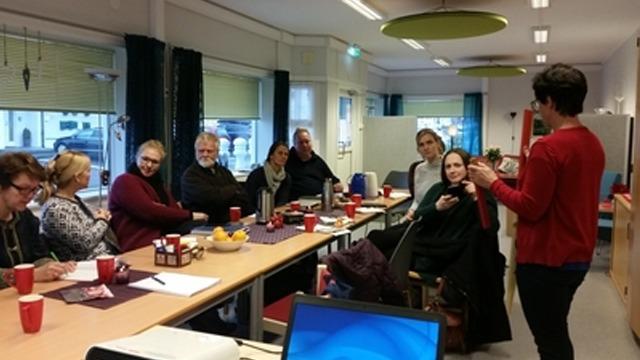 En gruppe mennesker sitter rundt et bord i et rom, med kopper og frukt på bordet, mens en person står og snakker.
