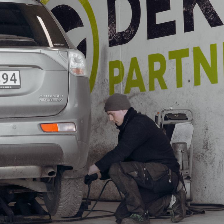En mekaniker jobber med å skifte dekk på en bil i et verksted med en stor logo på veggen.