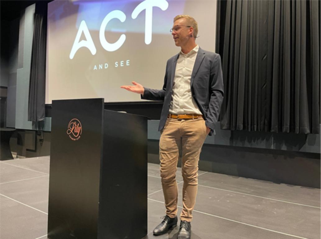 En mann står ved en talerstol på en scene med en projeksjon av teksten "ACT AND SEE" i bakgrunnen.