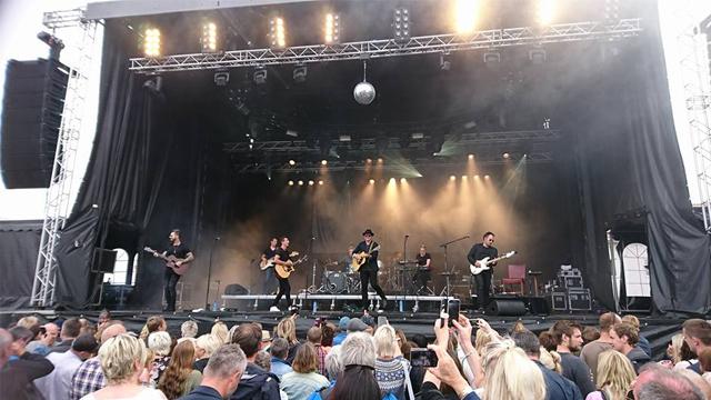 En konsert med et band som spiller på scenen, med publikum som ser på og danser i lyset fra scenen.