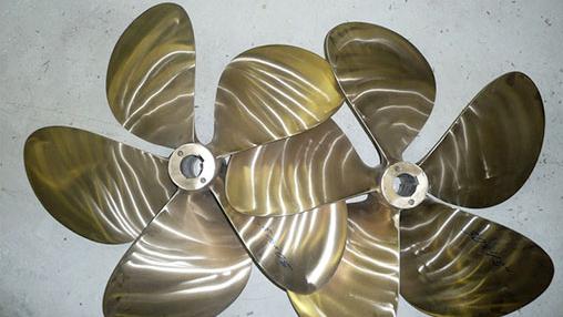To propellerblader i messing med fem blader, plassert på en grå overflate.