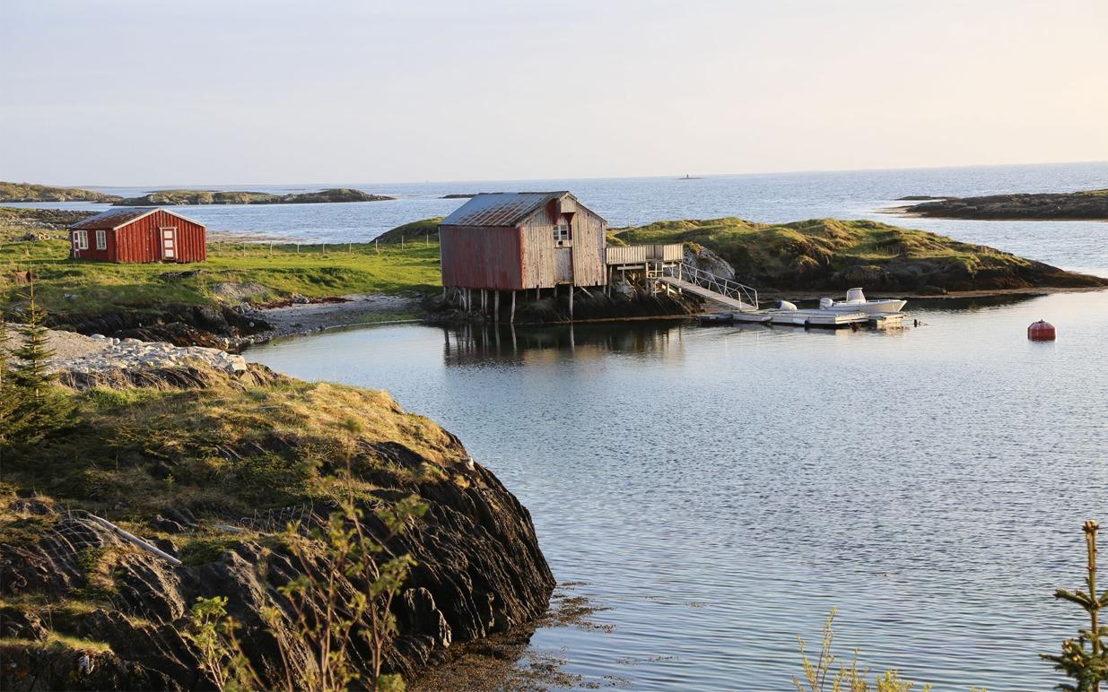 En rød hytte og et bryggehus ved en rolig bukt med små båter og steinete kystlinje.