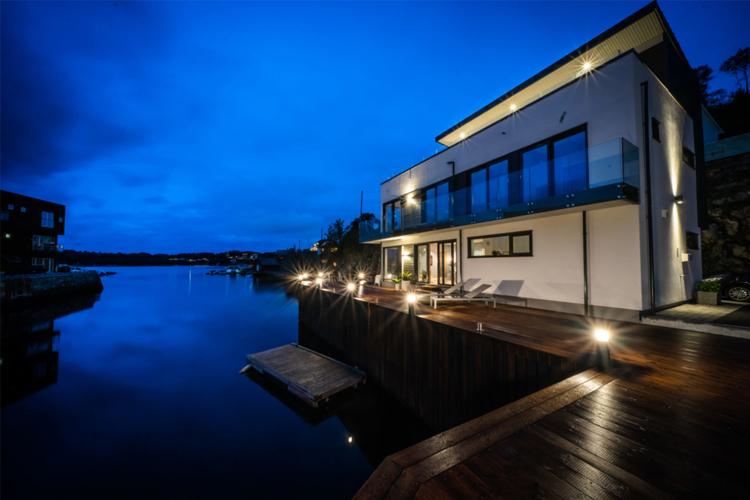 Moderne hus ved vannet om natten, med lys som reflekteres i vannet og en brygge i forgrunnen.