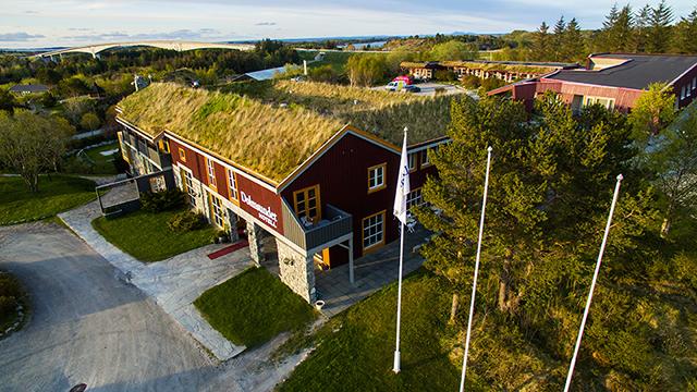 Rødt hus med grønt tak og hage, omgitt av trær og flaggstenger, beliggende i naturskjønt område.