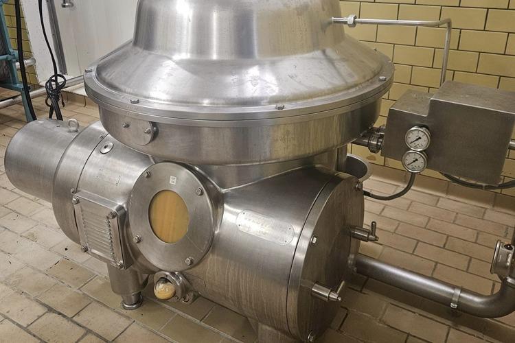 En stor, sølvfarget sentrifuge i rustfritt stål plassert på en flislagt gulv i et industrielt miljø.