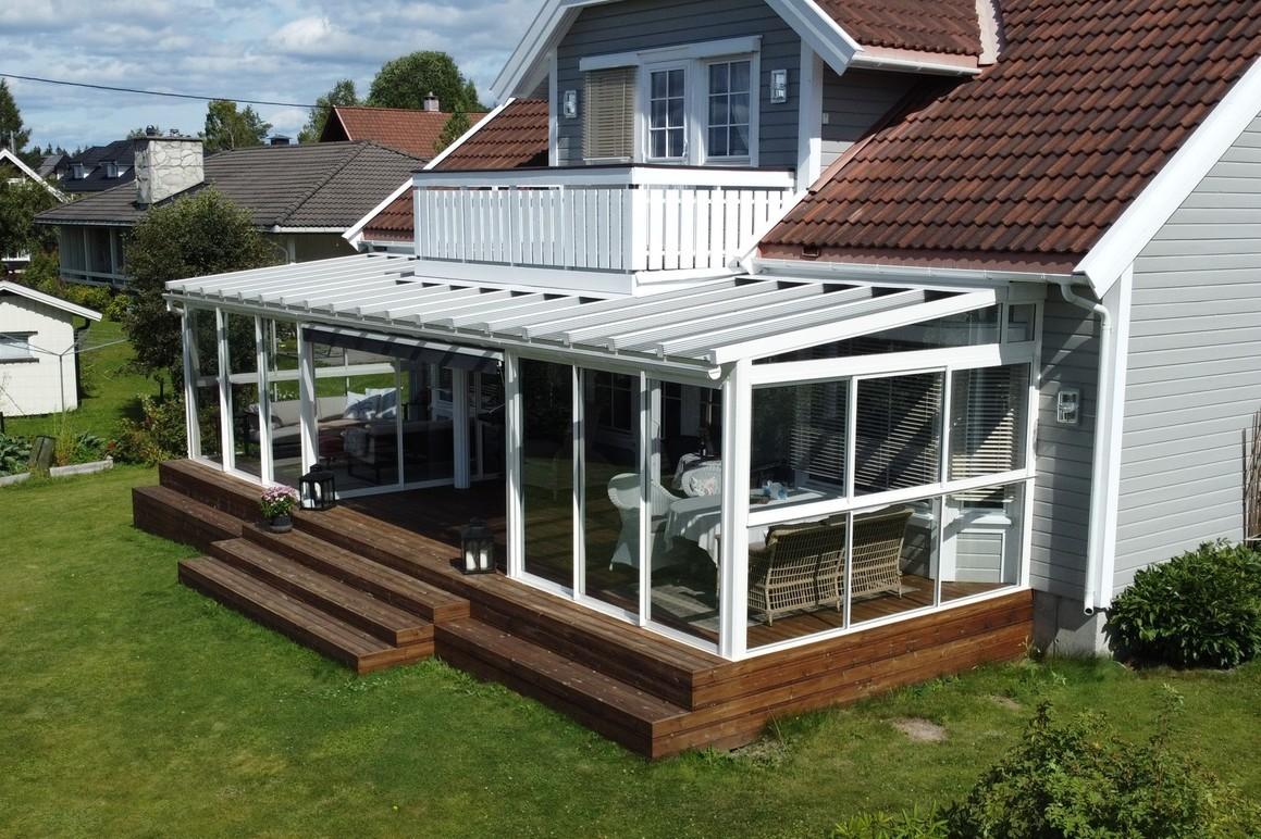 Moderne hus med stor terrasse og glassveranda, omgitt av grønt gress og trær.