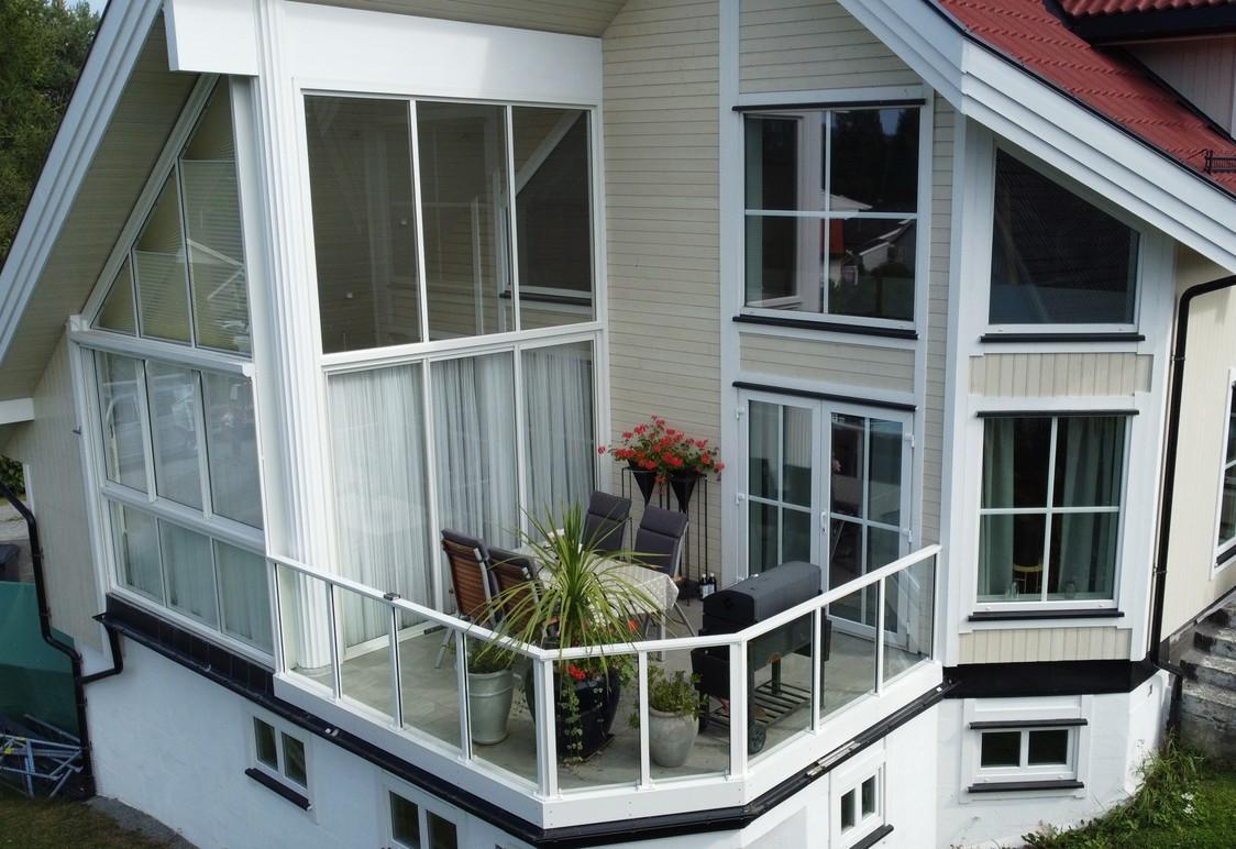 Moderne hus med stor balkong, glassvegger og møbler, omgitt av grønt landskap.