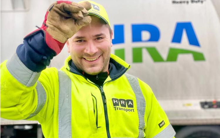 En smilende mann i gul arbeidsjakke og hansker, står foran en lastebil med HRA Transport-logo.