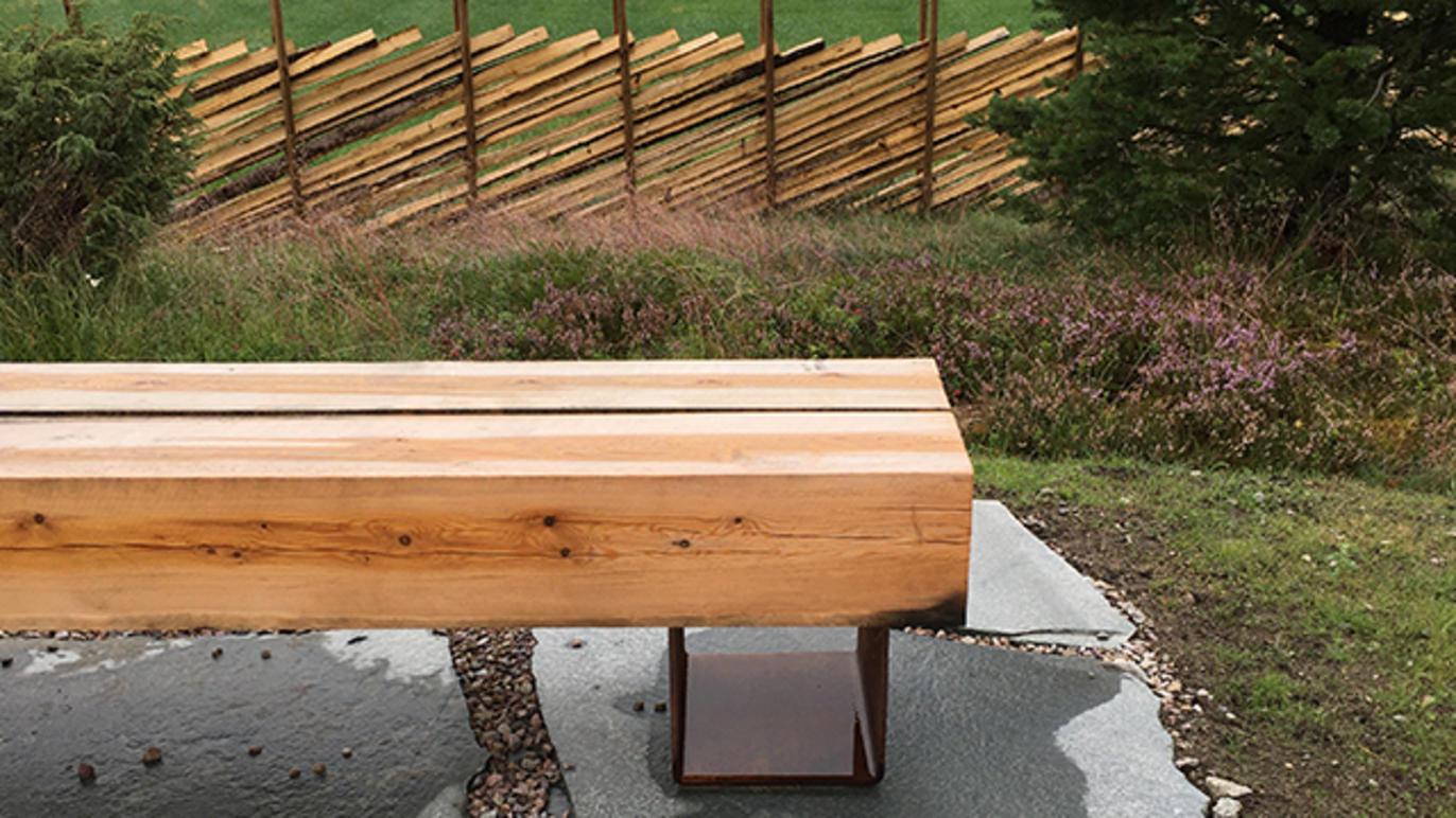En moderne benk laget av tre, plassert på en steinlagt overflate med en bakgrunn av trestammer.