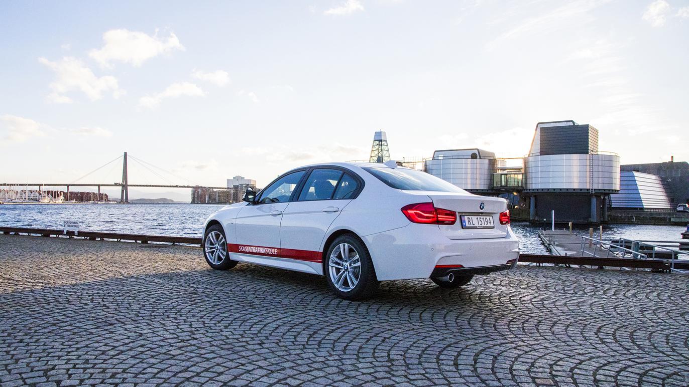 En hvit BMW sedan parkert ved vannkanten med moderne bygninger i bakgrunnen.