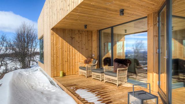 Moderne hytte med trevegg og terrasse, møblert med stoler, omgitt av snø og fjellandskap.