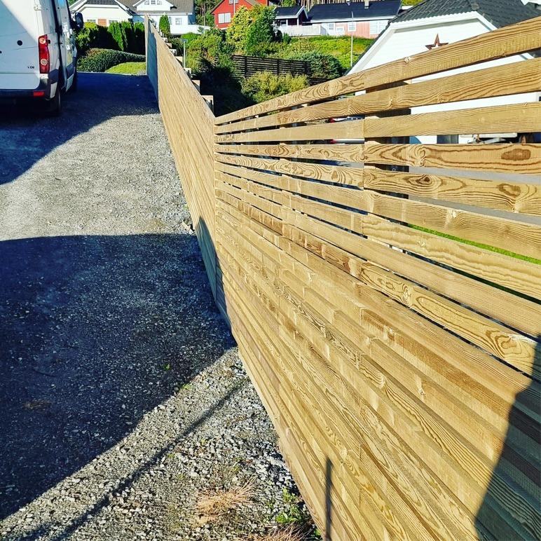 En moderne trevegg langs en grusvei med hus og hage i bakgrunnen.