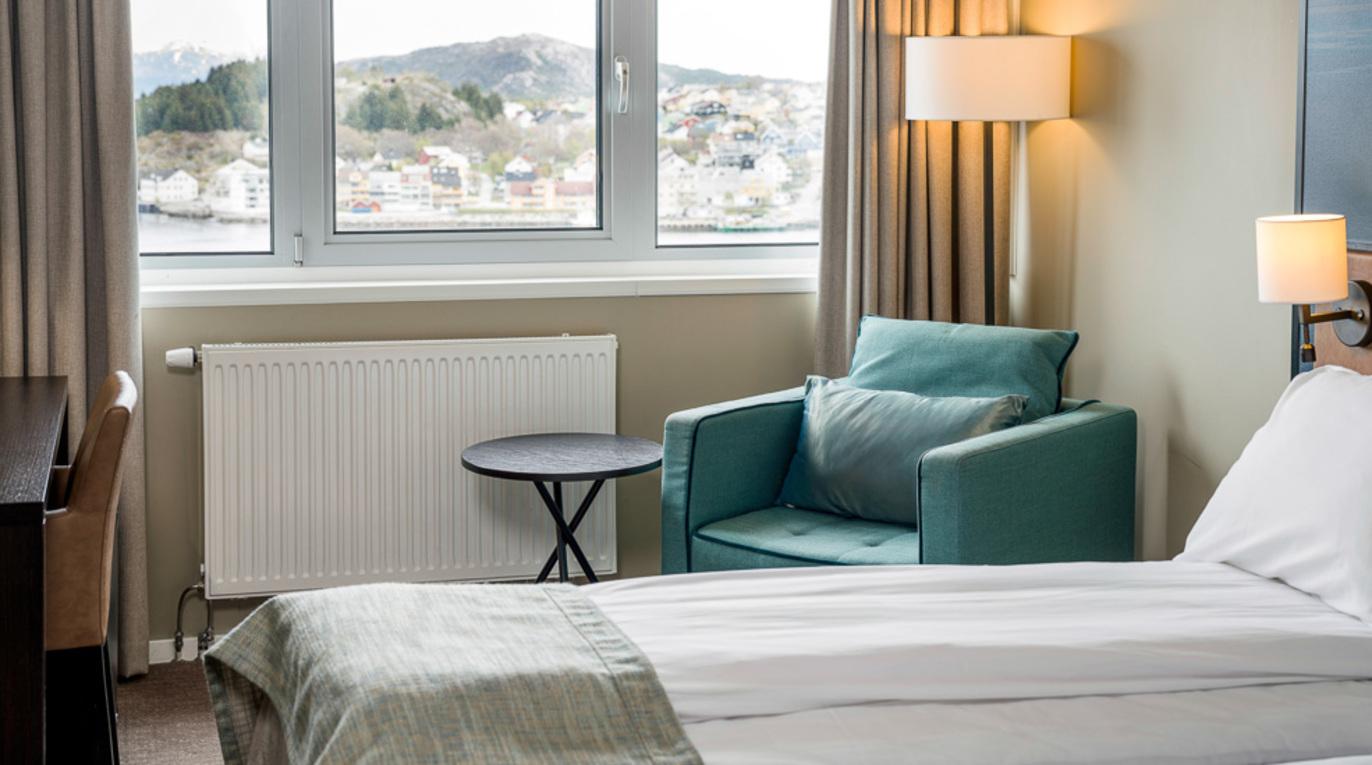 Moderne hotellrom med en seng, en grønn stol og vindu med utsikt over fjellene.