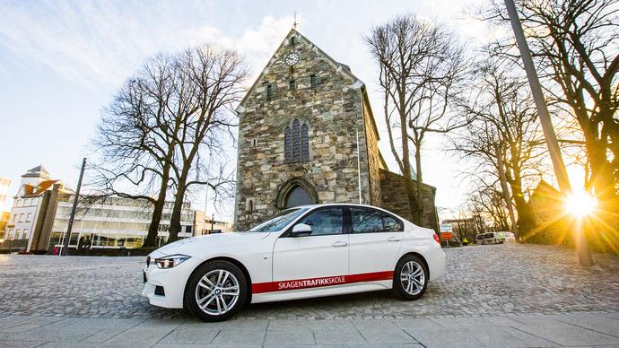 En hvit bil parkert foran en steinkirke med trær og solnedgang i bakgrunnen.