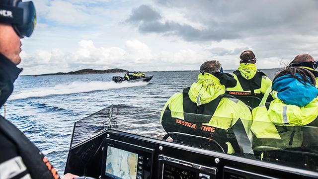 En gruppe mennesker i gule flytedresser på en båt, med en annen båt som kjører forbi i bakgrunnen.