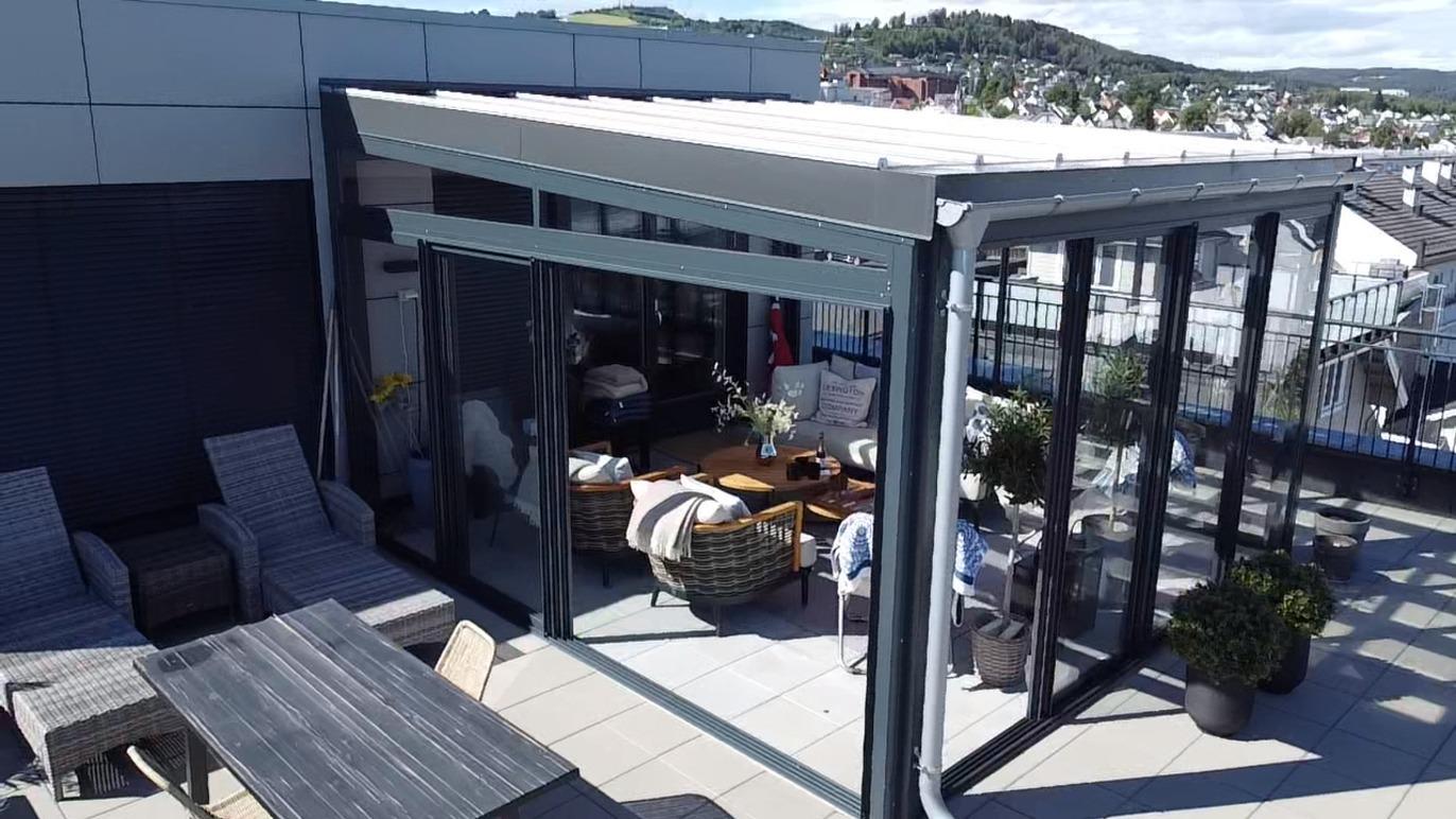 Moderne takterrasse med glassvegg, sittegruppe og spisebord, omgitt av planter og utsikt over landskapet.