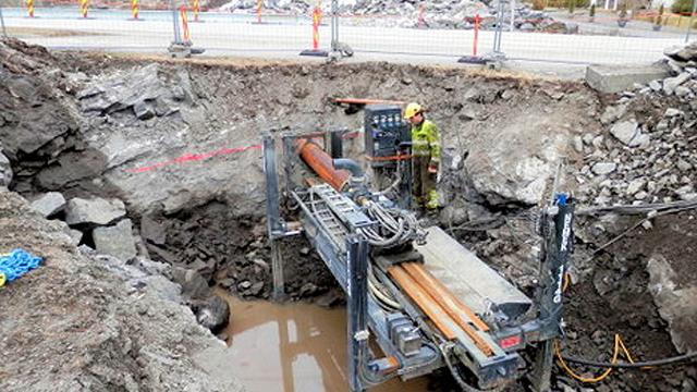 En graveringsmaskin i en dyp grøft med en arbeider iført verneutstyr og rør som ligger klart til installasjon.