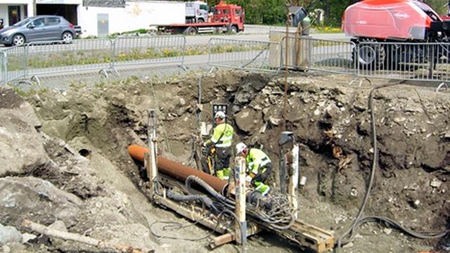 To personer i sikkerhetsutstyr arbeider i en grøft med rørleggerutstyr og maskiner i bakgrunnen.