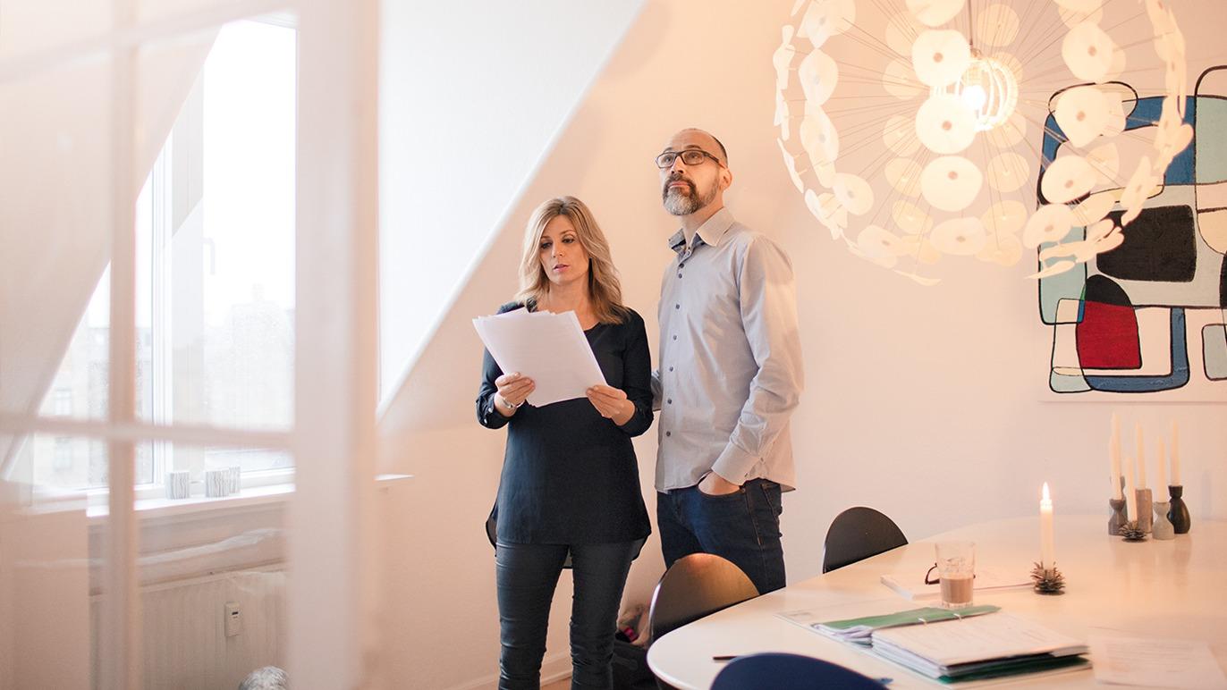 To personer står ved et bord i et lyst rom, ser på dokumenter og diskuterer. En moderne lampe henger over dem.