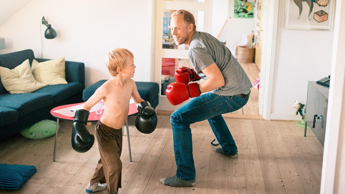 Far og sønn leker med boksehansker i stuen, med en sofa og bord i bakgrunnen.