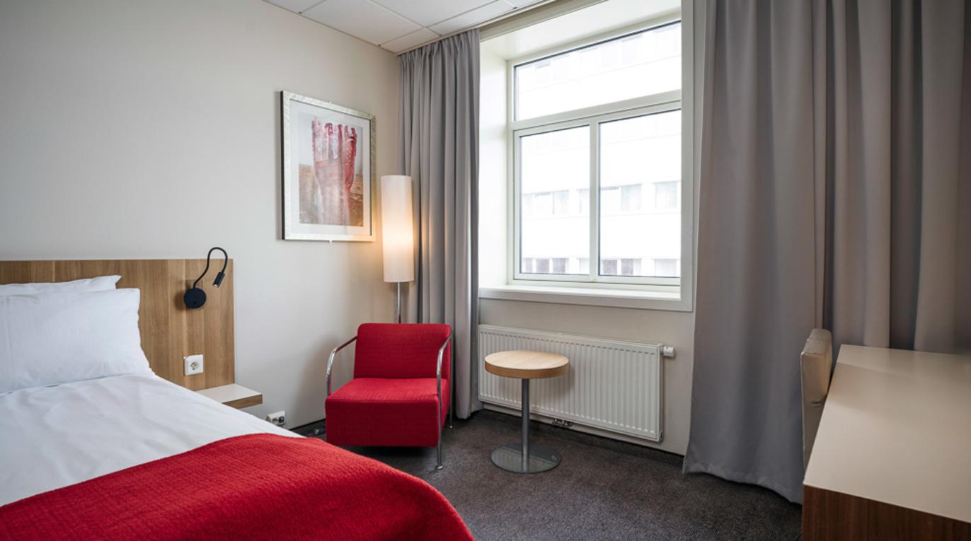 Moderne hotellrom med en seng, rød stol, bord og vindu med gardiner.