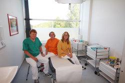Tre helsearbeidere i fargerike klær sitter på en behandlingsbenk i et lyst rom med medisinsk utstyr.