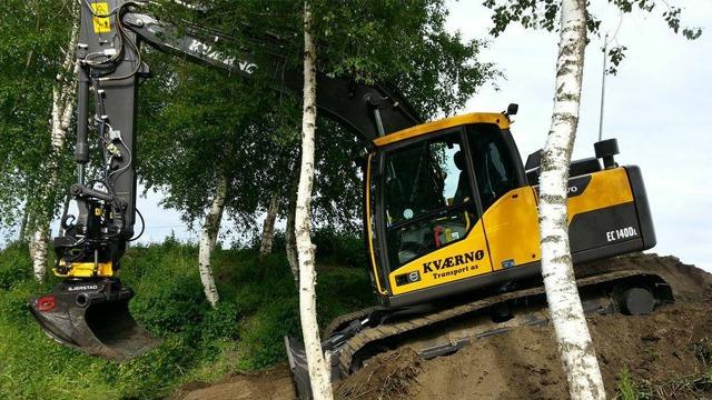 Gravemaskin i arbeid ved en skråning, omgitt av bjørketrær og grønt landskap.