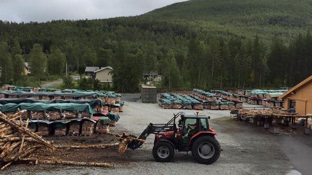 Traktor som laster ved på en gård med stablede vedkubber og skogkledd bakgrunn.