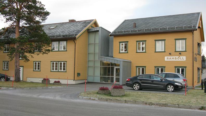 To bygninger med gult treverk og glassinngang, parkert bil foran, i en rolig gate.