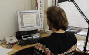 En person sitter ved en datamaskin med en skjerm, skriver på kontoret med papir og telefon i bakgrunnen.