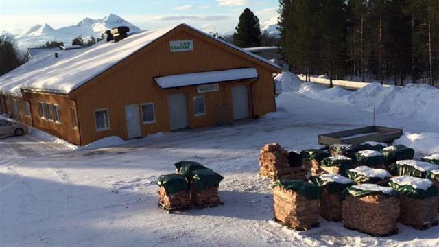 Gult bygg dekket av snø med høyballer stablet foran, omgitt av snødekte fjell i bakgrunnen.