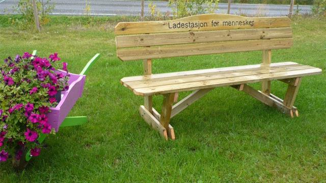 Benk i tre med teksten "Ladehaugen har minner" ved siden av en blomsterkasse med fargerike blomster.