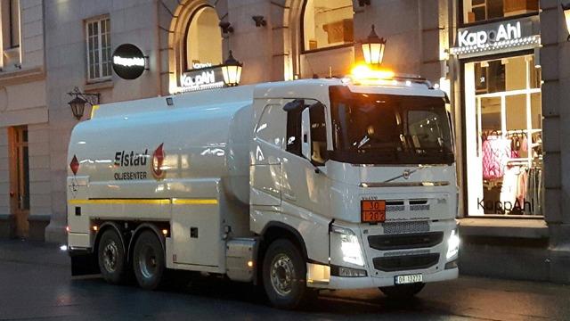En hvit tankbil med blinkende lys parkerer foran butikker i en gate om kvelden.