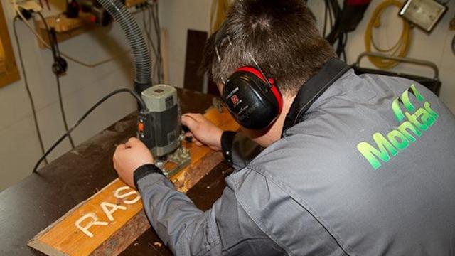 En person bruker en fresemaskin på en treplate med teksten "RAS" i en verkstedsetting.